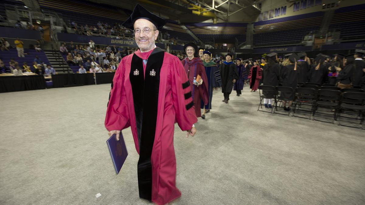 A man wearing graduation regalia leads a group of faculty out of a graduation ceremony.