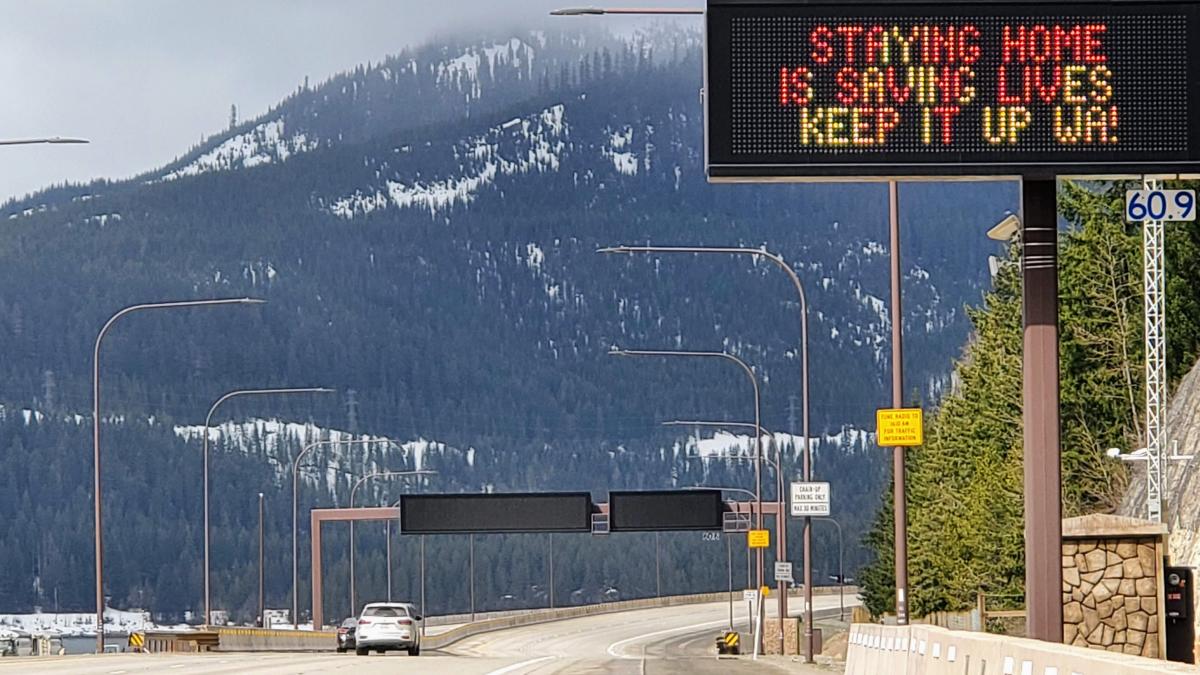 Two cars driving on otherwise empty Interstate 5 going over Snoqualmie Pass in Washington state, with roadside sign reading "STAYING HOME IS SAVING LIVES. KEEP IT UP WA!"