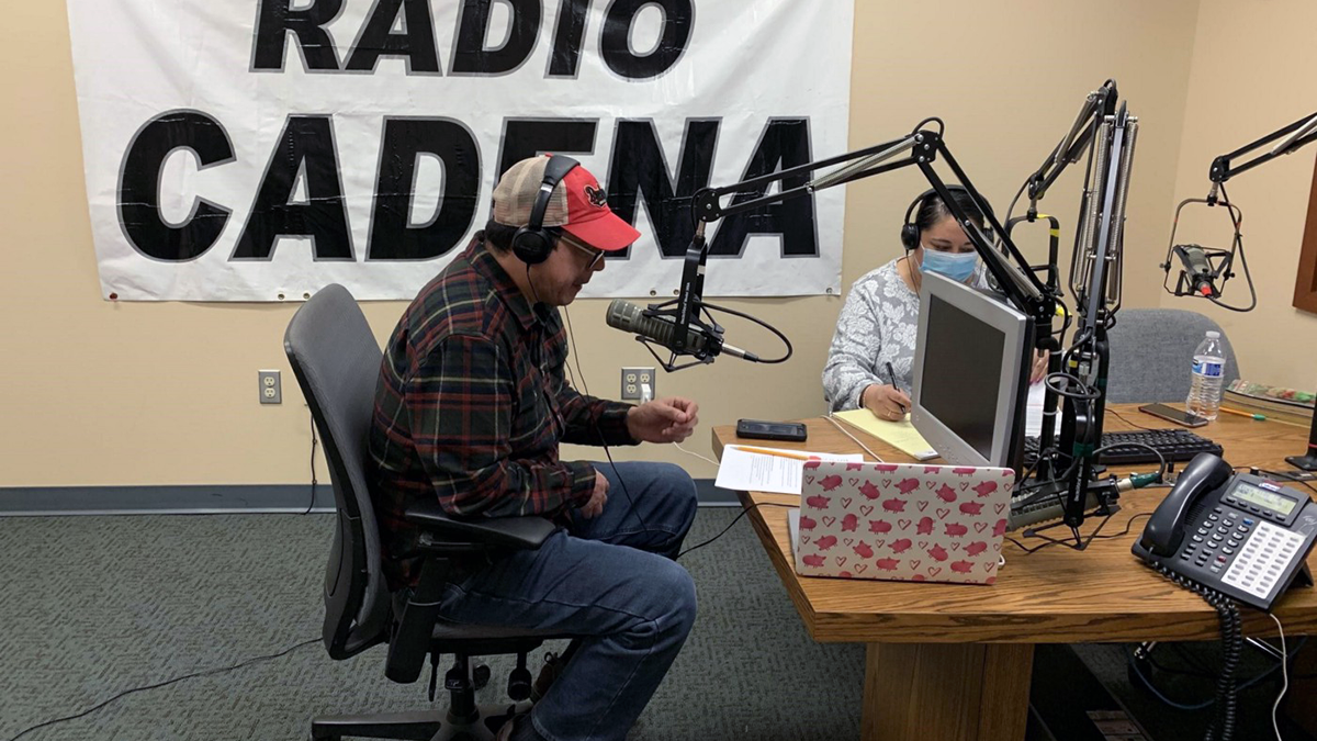 Two people sit in a radio booth at a table with microphones, with sign "RADIO CADENA" behind them.