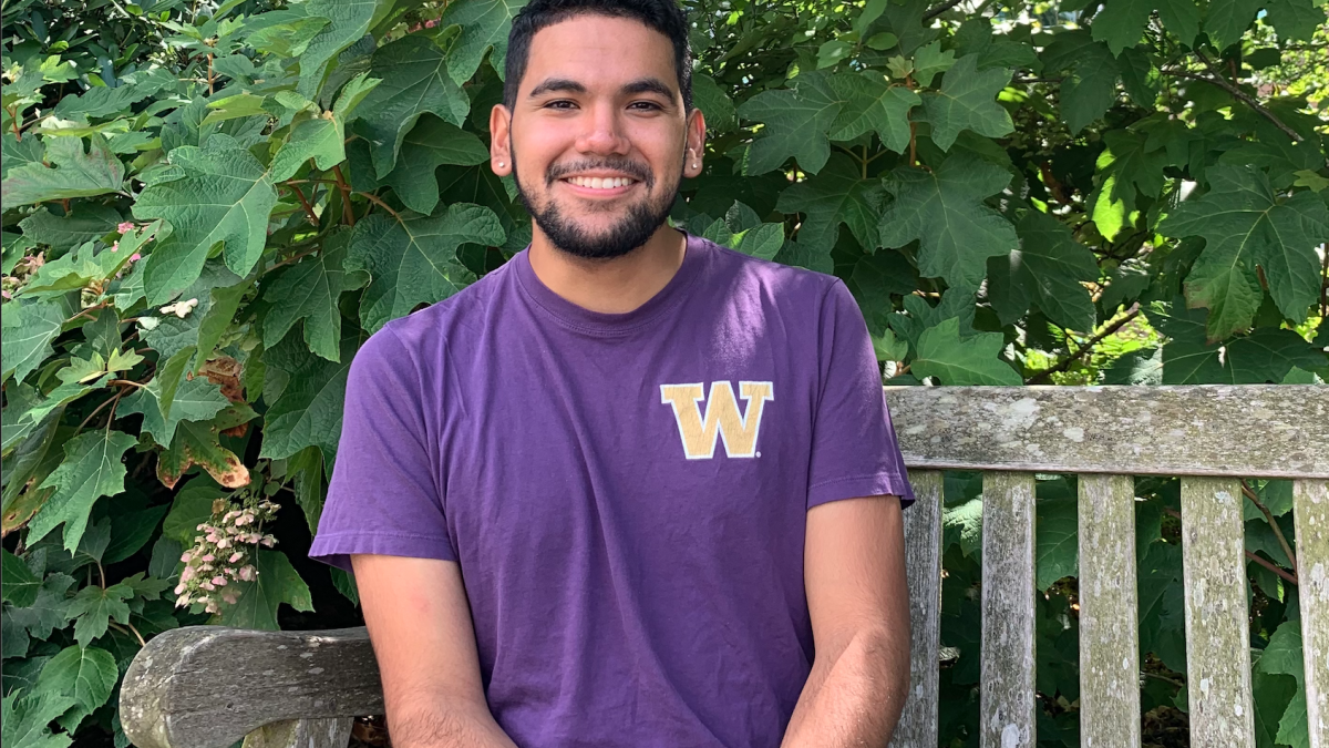 A man in a purple T-shirt sits on a bench in front of bushes.