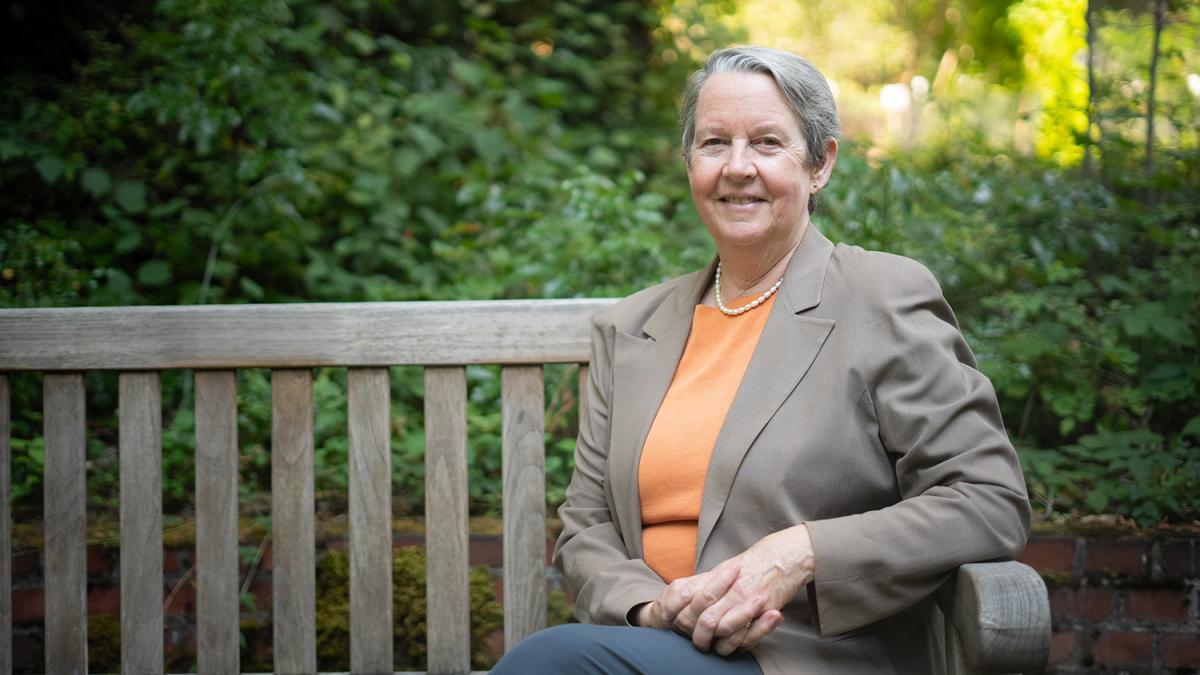 Lianne Sheppard sits on a wooden bench in front of green trees and shrubs. She wears a light orange shirt and light brown jacket with a pearl necklace and has her hands folded in her lap while smiling at the camera.