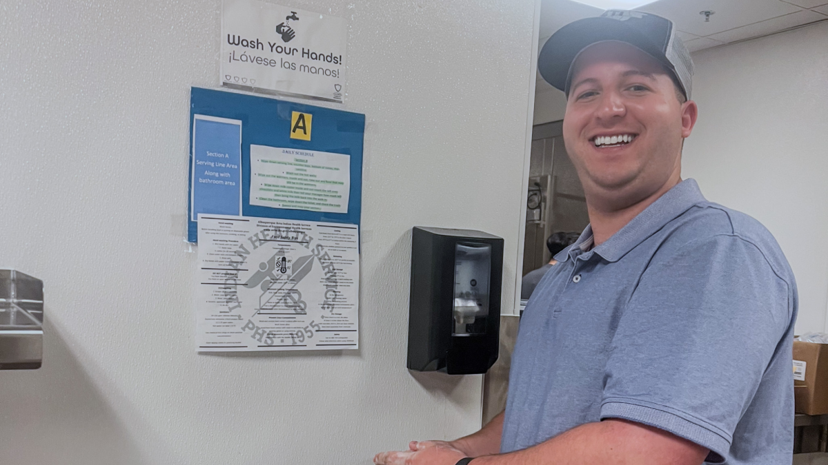 Jorge stands and washes his hands. On the wall there are several safety posters. One reads "Wash your hands Lavese las manos!" with a picture of a sink and hands. Jorge smiles at the camera and wears a blue shirt and hat.