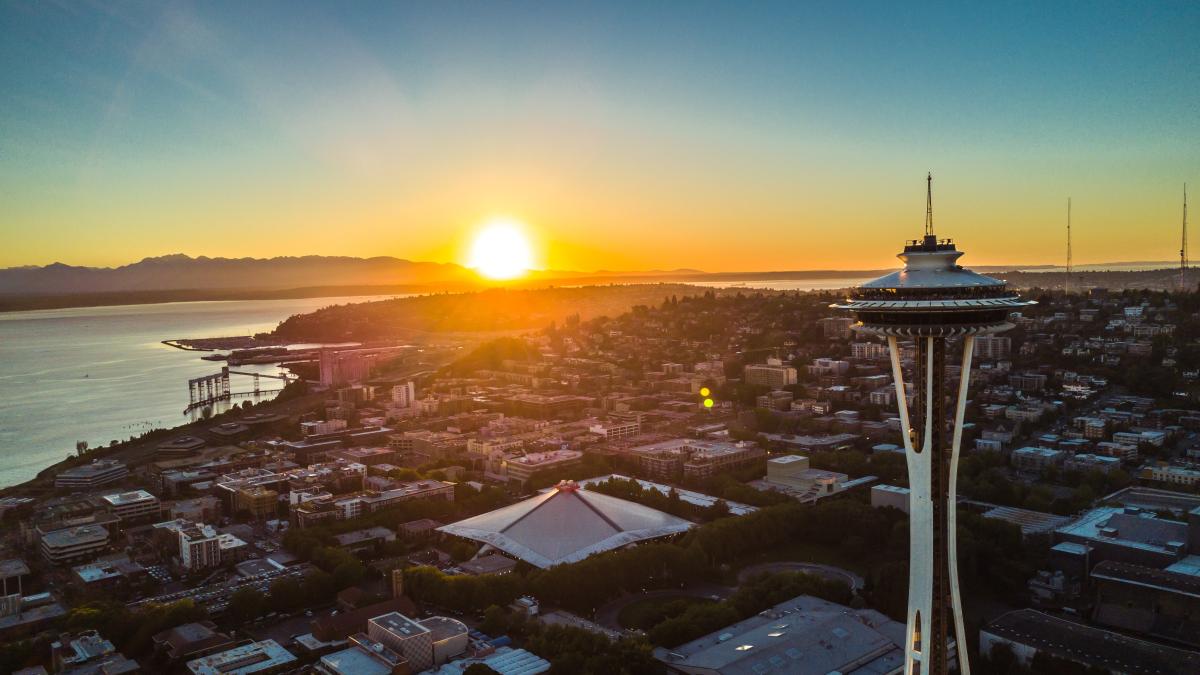 The Space Needle in the foreground with Seattle neighborhoods and a bright setting sun over Puget Sound and the Olympic Mountains in the background.