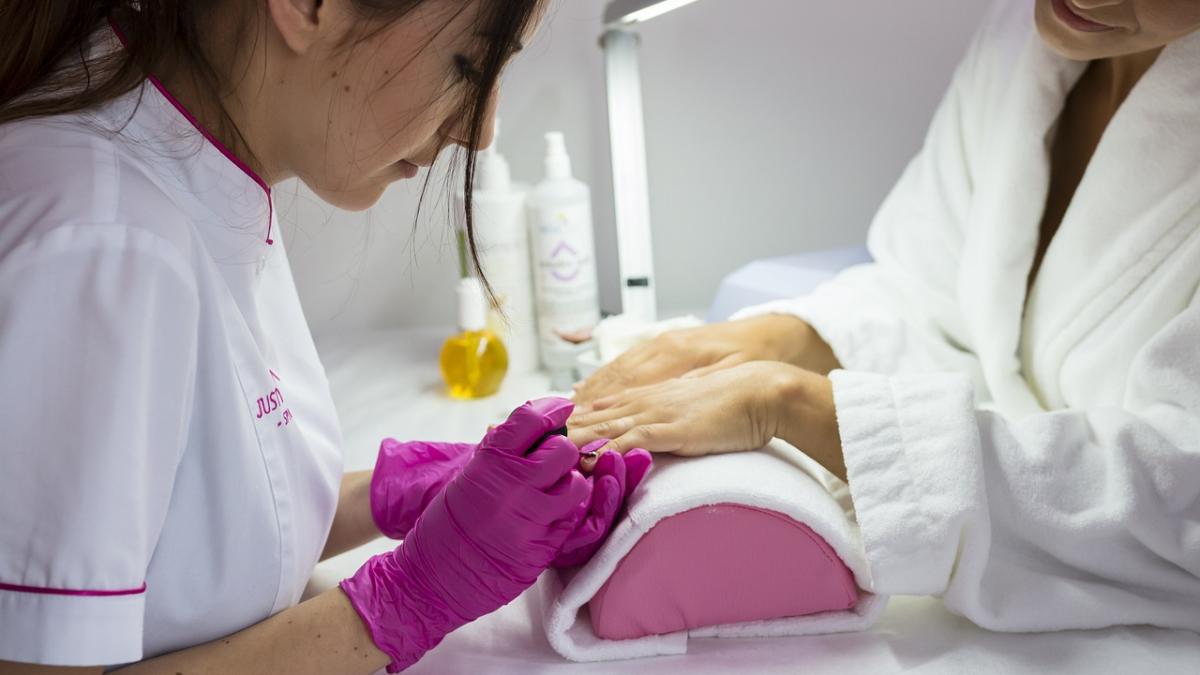 A nail technician wearing bright pink gloves and a white short sleeve salon jacket works on finger nails propped up on a cushion covered with a towel while the customer looks on. Nail products and a light are in the background.