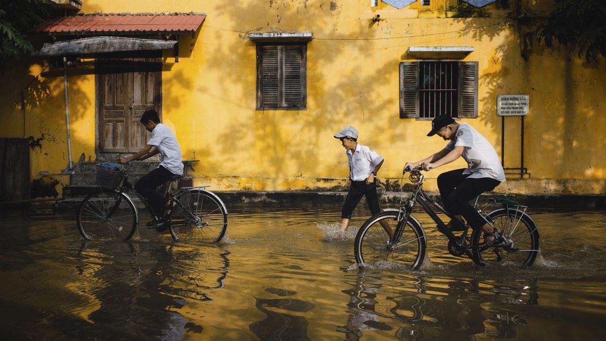 Two kids bike and one kid walks through flood water with a yellow shuttered building in the background.