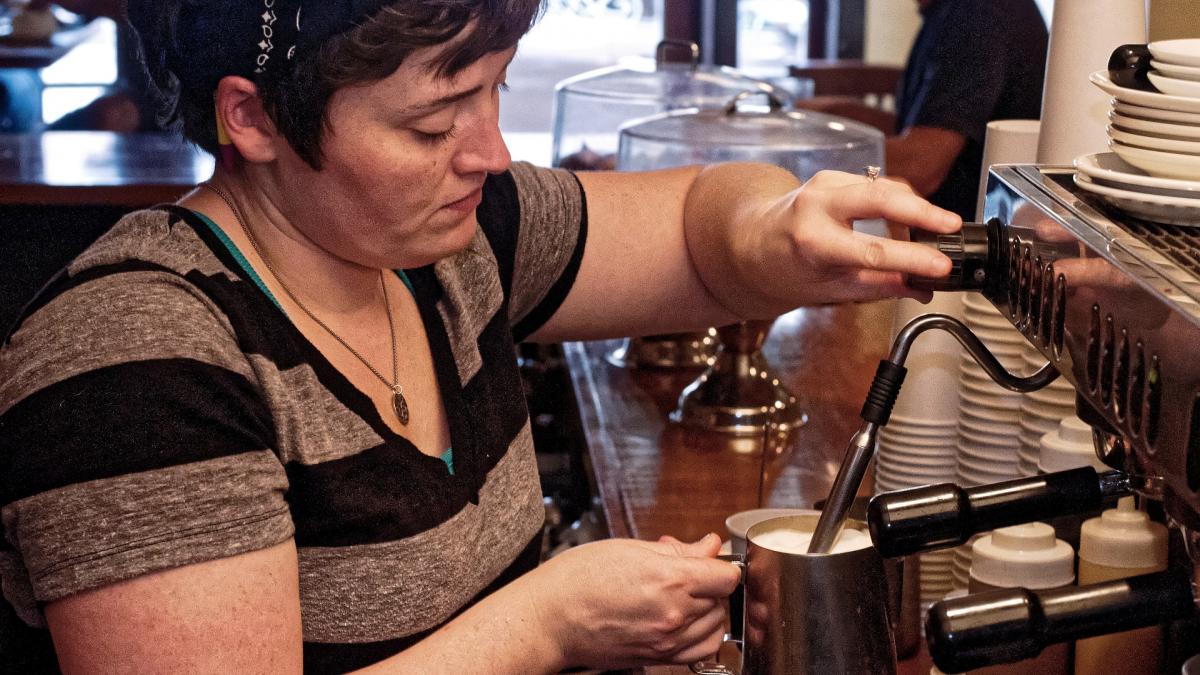 A woman in a coffee shop steams milk while preparing a coffee beverage.