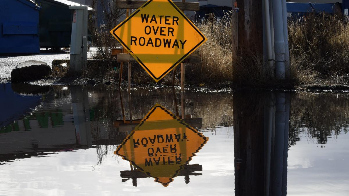 A yellow sign reading "water over roadway" is reflected in floodwaters.