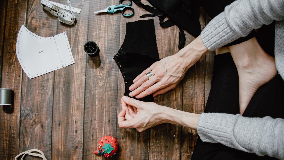 A person (only arms and legs shown) preparing a pattern for a handmade cloth face mask, sticking pins into it from a pincushion with thread, scissors and tape measure in the background.