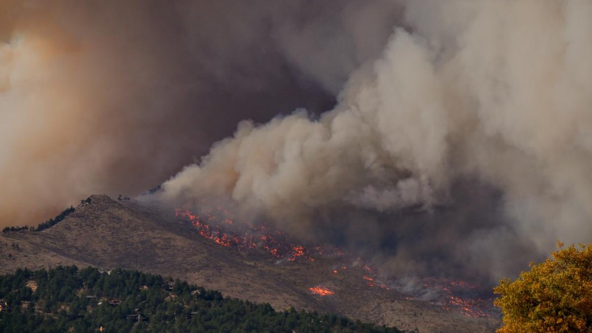 A smoke plume rises from a wildfire on a hillside near a group of houses.