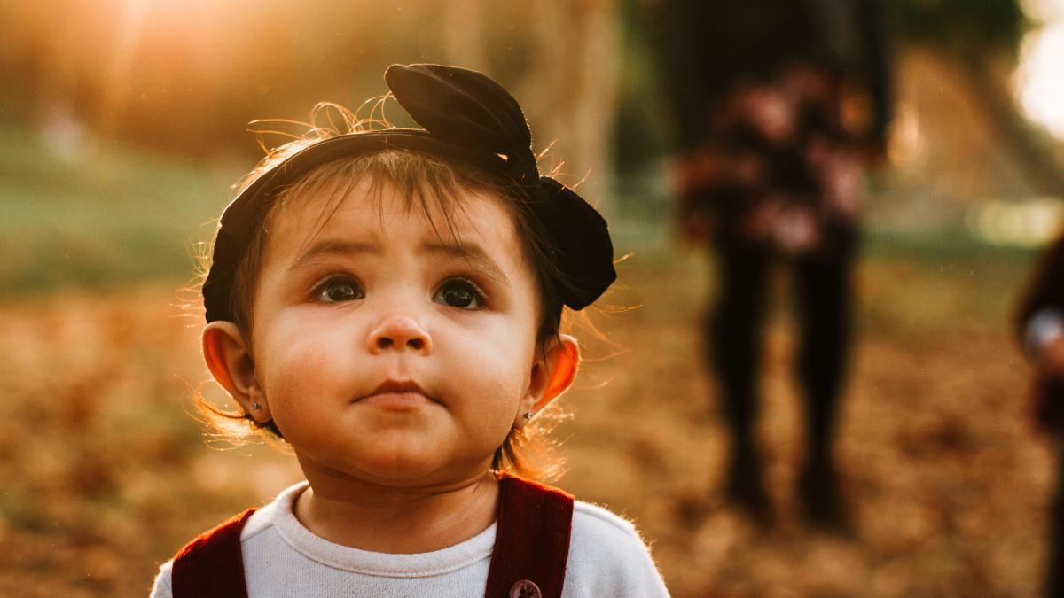 A toddler girl stands outside in a park with fallen leaves.