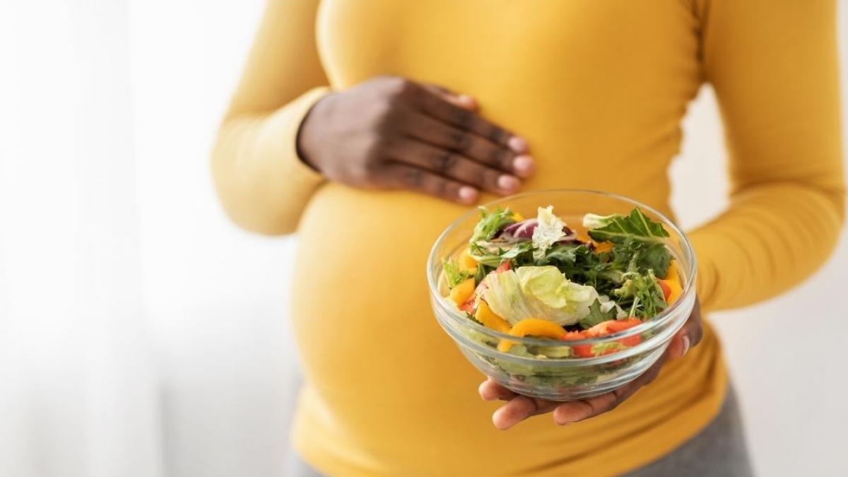 A woman dressed in a yellow shirt holds a bowl of leafy greens and other vegetables.
