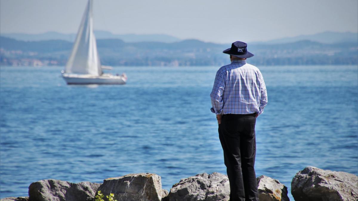 A man wearing a hat watches from shore as a sailboat passes by.