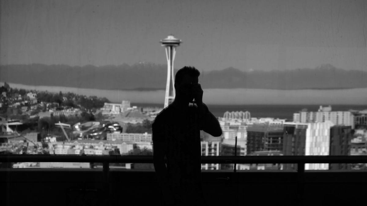 Man talking on phone in front of handrail with downtown seattle in the background. 