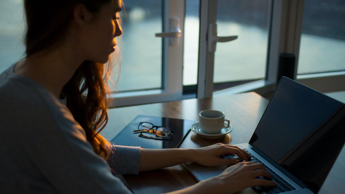 Woman looks down at laptop with her hands on the keys, indoors at a desk next to an open window.