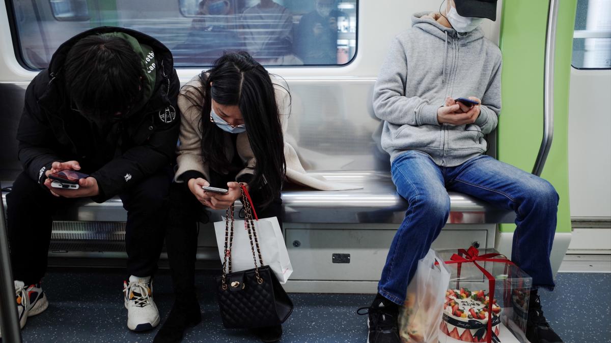 Three people sit on a subway wearing face masks and looking at their phones.