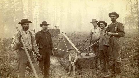 Black and white image of five loggers and a boy in Western Washington, W. T. The men are standing with axes in hand appearing to have just cut down a tree. The boy sits on the stump.