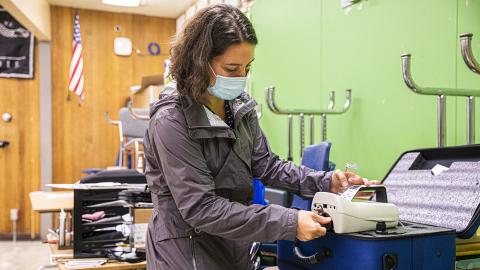 Woman with a surgical mask turns a dial on a portable air quality monitor in a classroom with a flag and desks in the background.