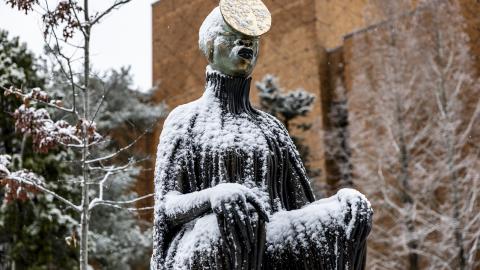 A bronze sculpture on the UW campus of a human figure seated with hands on knees, eyes closed  and a large flat circle on forehead. The sculpture is covered in light snow, with buildings, bare deciduous trees and an evergreen in the background..