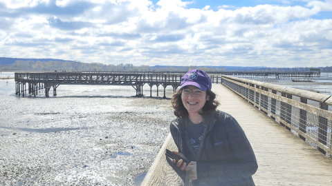 Cecilia Martindale stands smiling on a pier over a bay at low tide, wearing a purple UW hat.