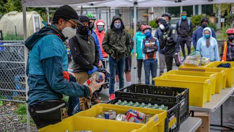 Bikers in masks stand around cartons of canned food under a tent.