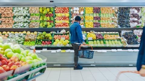 A person wearing jeans and a jean jacket stands in front of the produce section in a grocery store holding a grocery basket, with their back to the camera.