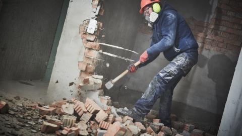 A worker wearing a respirator, helmet and ear protection demolishes a wall in a building with bricks on the floor below him.