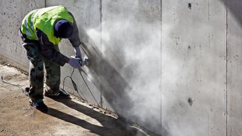 A worker in a safety vest facing away from the camera leans down to sand a concrete wall with dust flying into the air.