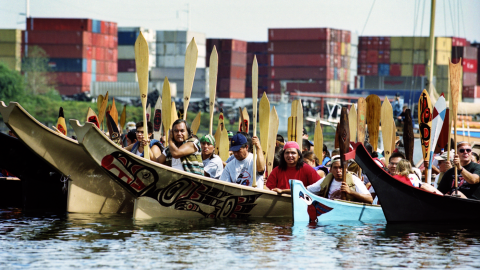 A group of people paddle Native boats on a river past a shipping terminal. 