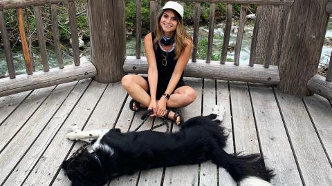 A woman wearing a hat sits on a wooden deck overlooking a river next to her black dog.