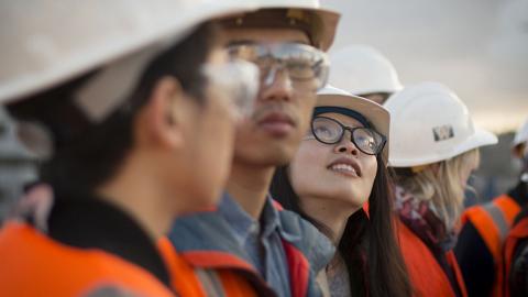 Student on a field trip to a construction site, looking up in a line of students. They are all wearing orange vests and white hard hats.