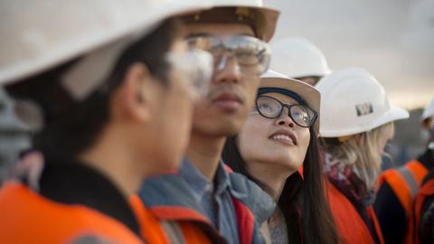 Student on a field trip to a construction site, looking up in a line of students.  They are all wearing orange vests and white hard hats.