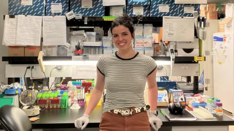 A woman in a striped T-shirt and wearing surgical gloves stands near a desk in a laboratory.