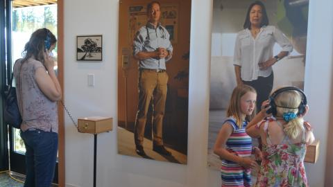 A woman wearing a headset stands at a listening station next to a tall portrait of a doctor, while two girls next to her look at a second tall portrait of a woman. 