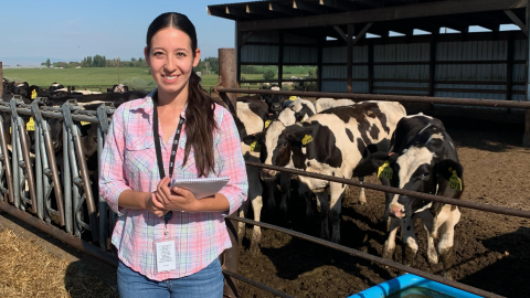 Diana Marquez stands in front of an enclosure of cows at a dairy farm with a notebook in her hands.