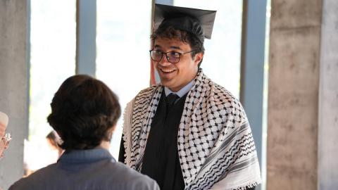 A student wears glasses, cap, gown and a keffiyeh covering his shoulders while talking with a person in the entryway of the Hans Rosling Center. 