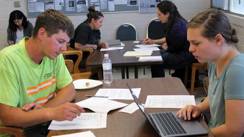 Students sit at tables interviewing construction workers.  In the foreground a female student with a laptop interviews a male construction worker with a packet of papers in front of him.