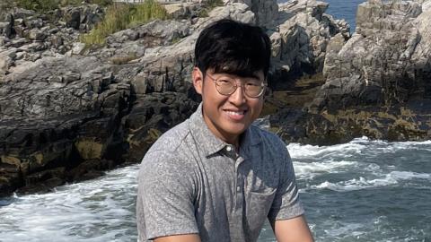 Kyle Kim smiles with ocean water and a rocky shoreline in the background.