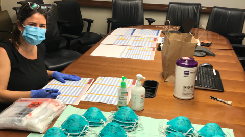 Woman with surgical mask and gloves on sits at a conference table with papers, sanitizer, wipes and N95 masks.