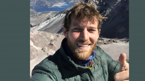 Photo of a man giving a thumbs up at the summit of a mountain.