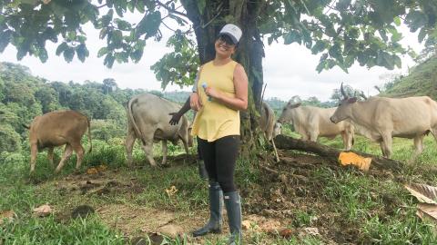 Viviana stands smiling in front of a tree and five cattle. She is holding a sampling tube and wearing glasses, a cap, a tanktop, yoga pants and tall rain boots.