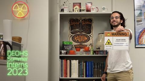 A man with dark hair and glasses stands in a radiation safety lab holding a sign that reads "Warning, Do not enter, Alignment in process. Class 4 laser"