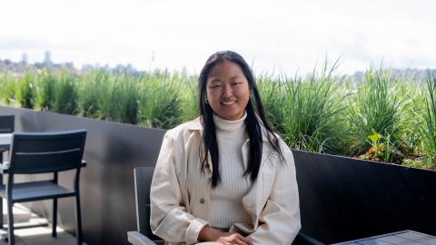 A young woman with long black hair and a white jacket sits at an outdoor table.