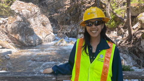 Kailee Sisounthone smiles wearing a hard hat, safety vest and sunglasses on a bridge over a river.