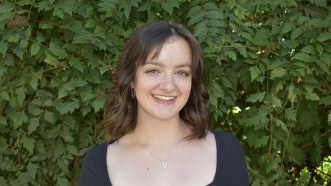 Cordy stands in front of greenery and smiles at the camera. Cordy wears a black shirt, silver necklace and silver earrings. 