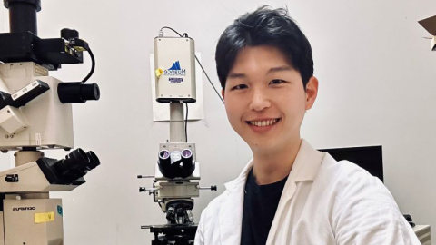 A man wearing a white lab coat smiles while sitting next to two microscopes.