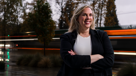 Portrait of a woman with long blond hair and a blue jacket standing outside as the blurred lights of passing traffic pass by.