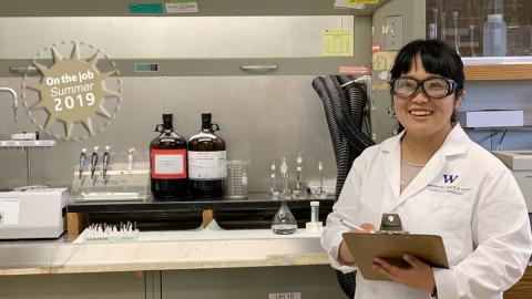 A young woman wearing a lab coat and safety goggles stands in a University of Washington laboratory.