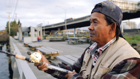 A man in a baseball cap and fishing vest stands with a fishing pole over a waterway.