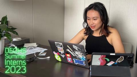 A young woman in a black tank top sits at a table looking at a computer.