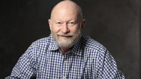 Richard (Rick) Gleason poses in front of a black background wearing a blue and white checked button up shirt, smiling a friendly smile at the camera.